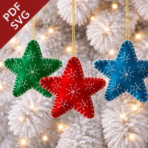 Könnte beinhalten: Drei sternförmige Filzornamente in Grün, Rot und Blau, jeweils mit weißer Schneeflockenstickerei und einem goldenen Faden zum Aufhängen. Die Ornamente sind vor einem verschneiten Weihnachtsbaum mit warmen Lichtern zu sehen.