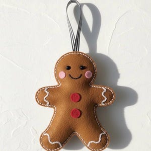 May include: A brown felt gingerbread man Christmas ornament with white stitching, pink cheeks, and red buttons. The ornament has a black and white striped ribbon for hanging. A festive holiday decoration.