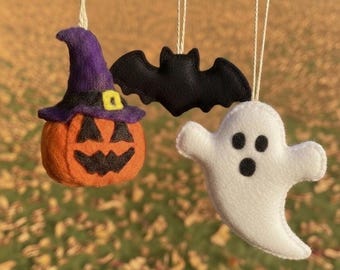May include: Three Halloween-themed felt ornaments hanging from strings. The ornaments include an orange pumpkin with a purple witch hat, a black bat, and a white ghost with black eyes. The background is blurred.