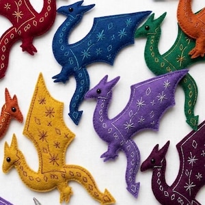 Op de afbeelding: Een verzameling kleurrijke vilten drakenornamenten in verschillende tinten, waaronder rood, blauw, groen, geel, oranje en paars. Elke draak heeft geborduurde details zoals sterren en diamanten, met een grillig, handgemaakt uiterlijk.