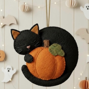 May include: A black cat ornament with orange ears and a pink nose, sleeping and hugging an orange pumpkin with a green leaf. The Halloween-themed decoration is made of felt and has a gold string for hanging.
