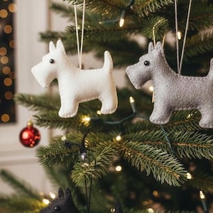 Puede incluir: Adornos navideños de fieltro con forma de terrier escocés en blanco, gris y negro. Los adornos de perro cuelgan de cuerdas blancas en un árbol de Navidad con luces y bolas rojas.
