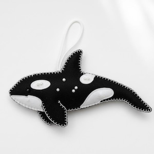 May include: A handmade felt orca ornament. The black and white killer whale has white accents and pearl embellishments. A white string is attached for hanging. The ornament is designed with a stitched outline.