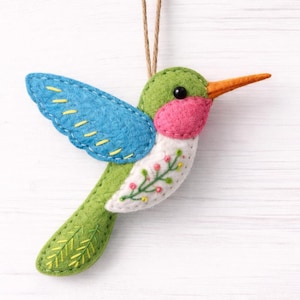 Puede incluir: Adorno de colibrí hecho a mano de fieltro, con cuerpo verde, alas azules y garganta rosa. El pájaro tiene un pico naranja y está decorado con costuras amarillas y detalles florales. Se adjunta una cuerda marrón para colgarlo.