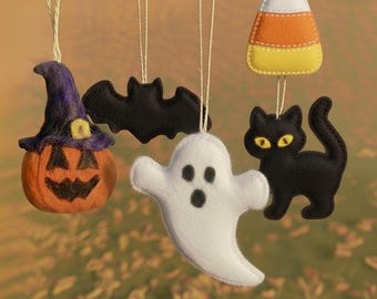 May include: A collection of Halloween-themed felt ornaments. Includes a jack-o'-lantern with a witch's hat, a black bat, a white ghost, a black cat with yellow eyes, and a candy corn. Each ornament is suspended by a string.