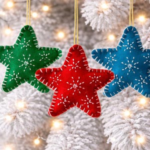 May include: Three felt star ornaments in green, red, and blue, each with white snowflake patterns and white stitching. The stars hang from gold strings against a backdrop of a snow-covered tree with twinkling lights.