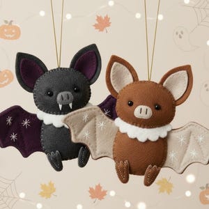 Könnte beinhalten: Zwei Fledermaus-Ornamente aus Filz, eines schwarz mit lila Flügeln und das andere braun mit beigefarbenen Flügeln, sind an goldenen Schnüren aufgehängt. Jede Fledermaus hat einen weißen Rüschenkragen und ist mit gestickten Details verziert. Der Hintergrund zeigt Halloween-Dekorationen.