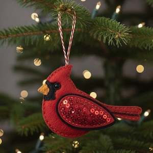 Peut inclure: Ornement de Noël en forme de cardinal rouge en feutre, avec des détails noirs et une aile ornée de paillettes. L'oiseau a un bec jaune et une ficelle de suspension rouge, blanche et argentée. Accroché à un sapin de Noël vert.