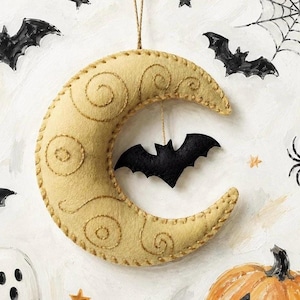 May include: A crescent moon ornament made of beige felt with decorative gold thread stitching in swirl patterns. A small black felt bat hangs from the centre of the moon by a thin string. The ornament is suspended by a gold cord for hanging.