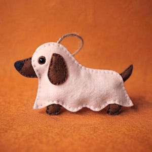 Puede incluir: Un adorno de fieltro con forma de perro salchicha que muestra la cabeza y las patas marrones asomando bajo un disfraz de fantasma rosa pálido. La funda de fantasma tiene un borde inferior festoneado y un pequeño lazo para colgar.