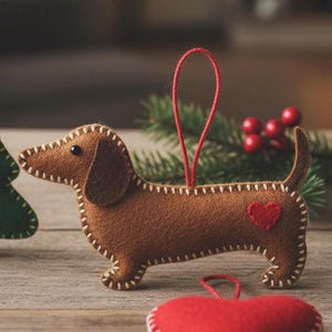 Può includere: Un ornamento a forma di bassotto in feltro marrone con un cuore rosso e dettagli cuciti bianchi. Il cane ha un anello rosso per appenderlo ed è ambientato su uno sfondo sfocato con verde e bacche rosse. Un ornamento a forma di cuore rosso è in primo piano.