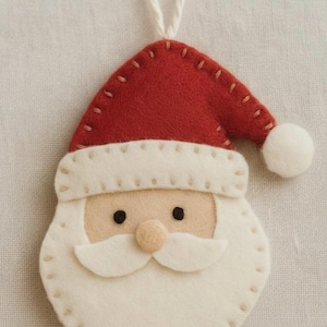 May include: A festive Christmas decoration featuring a felt Santa Claus face. The ornament has a red hat with a white trim and a white beard and moustache. It has black eyes and a small pink nose, with a white string for hanging.