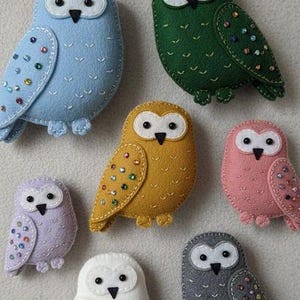 May include: A selection of handmade felt owl decorations in shades of blue, green, yellow, pink, purple, white, and grey. Each owl features a white face, black eyes, and decorative embellishments on the wings. These charming decorations are ideal for a nursery or home decor.