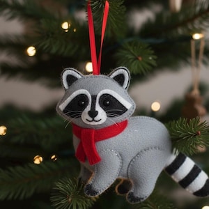 Puede incluir: Adorno navideño de fieltro gris con forma de mapache, con bufanda roja y cola a rayas blancas y negras. El adorno tiene una cinta roja para colgar y está en un árbol de Navidad.
