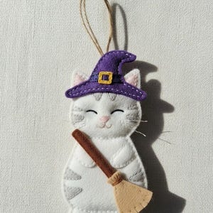 以下が含まれることがあります： 灰色の縞模様が入った白いフェルト猫のオーナメント。紫色の魔女の帽子と金のバックルをかぶっています。猫は茶色のほうきを持っており、吊るすための紐が付いています。かわいいハロウィーンの装飾。