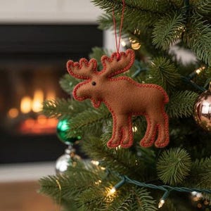 May include: A brown felt moose Christmas ornament with red stitching and a red hanging string. The moose is hanging on a green Christmas tree decorated with ornaments and lights. A fireplace is visible in the background.