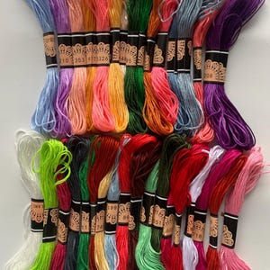 Può includere: Una collezione di gomitoli di filo da ricamo colorati in varie tonalità di rosa, rosso, verde, blu, giallo e viola. Ogni gomitolo è avvolto con un'etichetta nera con testo bianco e un design decorativo. Ideale per progetti di ricamo e punto croce.