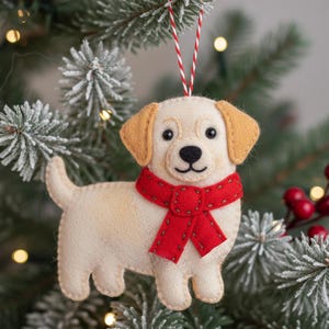 Puede incluir: Adorno navideño de fieltro con forma de perro, con una bufanda roja y orejas beige. El perro tiene nariz y ojos negros. Cuelga de un cordón rojo y blanco en un árbol de Navidad.