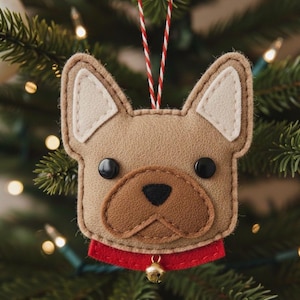 Op de afbeelding: Een kerstboomversiering van vilt in de vorm van een Franse bulldogkop. De lichtbruine versiering heeft crèmekleurige oordetails, zwarte knoopogen en een bruine neus. Het heeft een rode kraag met een kleine gouden bel en een rood-wit touwtje om op te hangen.