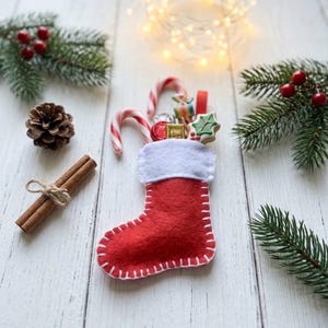 May include: A small red felt Christmas stocking with a white cuff and white stitched edges, filled with candy canes, a small wrapped gift, and a holly-shaped cookie. The stocking is surrounded by pine branches, a pinecone, and cinnamon sticks.