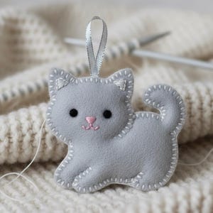May include: A gray felt cat ornament with a silver ribbon for hanging. The cat has black eyes, a pink nose, and white stitched details. The ornament is on a white knitted surface.