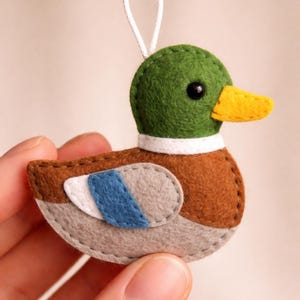 Puede incluir: Adorno de pato de fieltro hecho a mano. El pato tiene una cabeza verde, un cuerpo marrón y detalles de alas grises y azules. Presenta un pico amarillo, un ojo negro y un cuello blanco. Se adjunta una cuerda blanca para colgar.