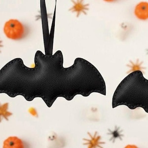 Op de afbeelding: Zwarte vleermuisvormige Halloween-ornamenten met gestikte randen hangen aan zwarte linten. De achtergrond toont kleine oranje pompoenen, stervormige decoraties en spookvormige snoepjes.