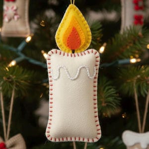 May include: A felt Christmas ornament shaped like a candle. The candle is off-white with a red stitched border and a white, wavy, dripped wax design. The flame is yellow and orange. The ornament hangs from a gold string.