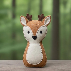 May include: A handmade felt deer plushie with brown, white, and black accents. The deer has a stitched design, small antlers, and a white belly. The plushie is standing upright against a blurred green background.