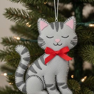 Könnte beinhalten: Graue Filzkatzen-Ornament mit schwarzen Streifen, rosa Innenohren und einer roten Schleife. Die Katze hängt an einer weißen Schnur vor einem Weihnachtsbaum. Eine charmante Weihnachtsdekoration.