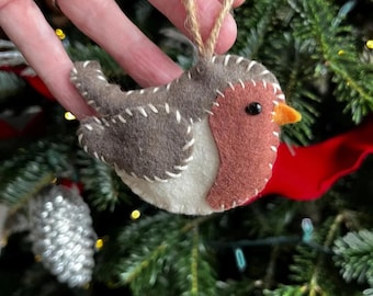 Puede incluir: Un adorno de fieltro con forma de pájaro que presenta un dorso marrón, vientre color crema y pecho rojo. El pájaro tiene un pequeño ojo de cuenta negra, un pico de fieltro naranja y costuras blancas en los bordes. Incluye un lazo de yute.