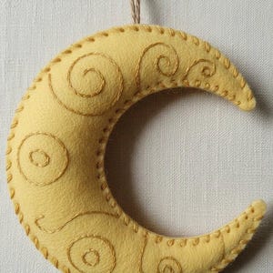 Puede incluir: Adorno artesanal en forma de media luna de fieltro amarillo. Presenta espirales bordadas en dorado y un borde cosido. Incluye un lazo de cuerda para colgar. Un elemento decorativo para el hogar, de unos 15 cm de largo.