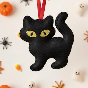 Könnte beinhalten: Schwarzer Filz-Katzen-Anhänger mit gelben Augen und rotem Band zum Aufhängen. Umgeben von Halloween-Dekorationen wie Kürbissen, Spinnen, Geistern und Zuckermais.