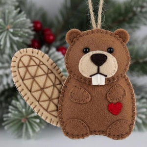 Pode incluir: Enfeite de feltro castanho em forma de castor com cauda castanho-claro e coração vermelho. O castor tem olhos pretos, nariz bege e dentes brancos. Uma argola de barbante é presa para pendurar. O enfeite está sobre ramos de sempre-vivas e bagas vermelhas.