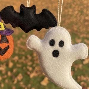 Op de afbeelding: Handgemaakte Halloween-ornamenten. Een witte vilten geest met zwarte ogen en mond, een zwarte vilten vleermuis en een oranje pompoen met een paarse hoed. Elk ornament is opgehangen aan een dun, natuurlijk gekleurd touwtje.