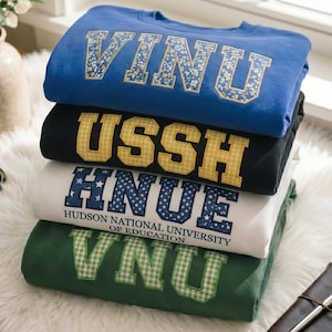 May include: A stack of four sweatshirts in blue, black, white, and green. Each sweatshirt has a different text design in a contrasting color. The blue sweatshirt has floral lettering, the black has yellow gingham lettering, the white has blue star lettering, and the green has green gingham lettering.