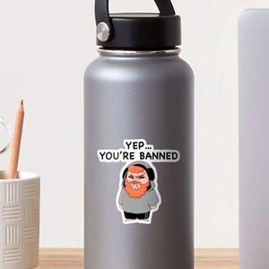 May include: A gray water bottle with a black lid. A sticker on the bottle features a cartoon character with a red beard and the text "Yep... You're Banned."