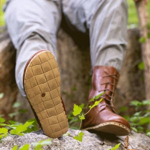 Op de afbeelding: Bruine leren wandelschoenen met veters en een textuurzool. De laarzen worden gedragen met een grijze broek en een lichtgroen shirt. De foto is buiten genomen, met een vage groene achtergrond.