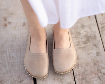 Puede incluir: Un par de zapatos sin cordones de cuero beige con punta redondeada y costuras blancas visibles a lo largo de la suela. Los zapatos presentan un acabado liso y mate con una abertura baja, usados con una prenda blanca sobre los tobillos.