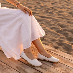 Op de afbeelding: Witte leren ballerina's met een ronde neus en een gestikte, lichtbruine zool. De schoenen worden gedragen met een witte jurk op een houten oppervlak, met een zandstrand op de achtergrond.
