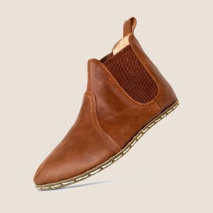 Könnte beinhalten: Ein brauner Chelsea-Stiefel aus Leder mit spitzer Zehenpartie und elastischem Seiteneinsatz. Der Stiefel hat eine Zuglasche am Rücken und eine genähte Sohle. Das Design ist minimalistisch und stilvoll, passend für verschiedene Outfits.