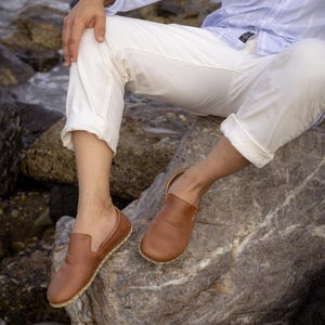 May include: Brown leather slip-on shoes with a rounded toe and a stitched sole. The shoes are shown on a person sitting on a rock, wearing white pants and a light blue shirt.