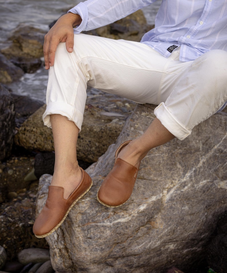 May include: Brown leather slip-on shoes with a rounded toe and a stitched sole. The shoes are shown on a person sitting on a rock, wearing white pants and a light blue shirt.