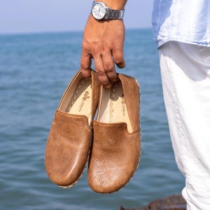 May include: A pair of brown leather slip-on shoes with a textured finish. The shoes have a rounded toe and a stitched sole. The background shows a body of water.