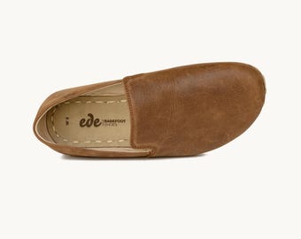 May include: A brown leather slip-on shoe with a rounded toe. The shoe has a stitched sole and a textured upper. The brand name "ede BAREFOOT SHOES" is printed on the insole. The shoe is a size W6.