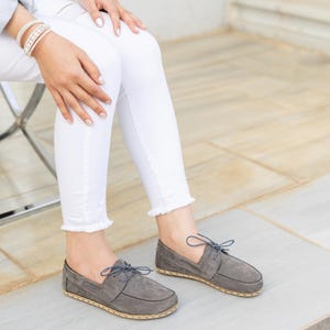 Könnte beinhalten: Ein Paar Bootsschuhe aus grauem Wildleder mit flacher Silhouette, dünnen grauen Schnürsenkeln und einem sichtbaren, hellen, genähten Rahmen entlang der Sohle. Die Schuhe haben eine abgerundete Zehenpartie und eine flache Konstruktion.