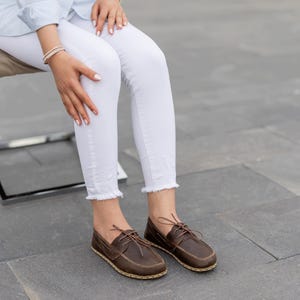 May include: Brown leather boat shoes with laces. The shoes have a stitched sole and are worn with white jeans. The shoes are a dark brown color.