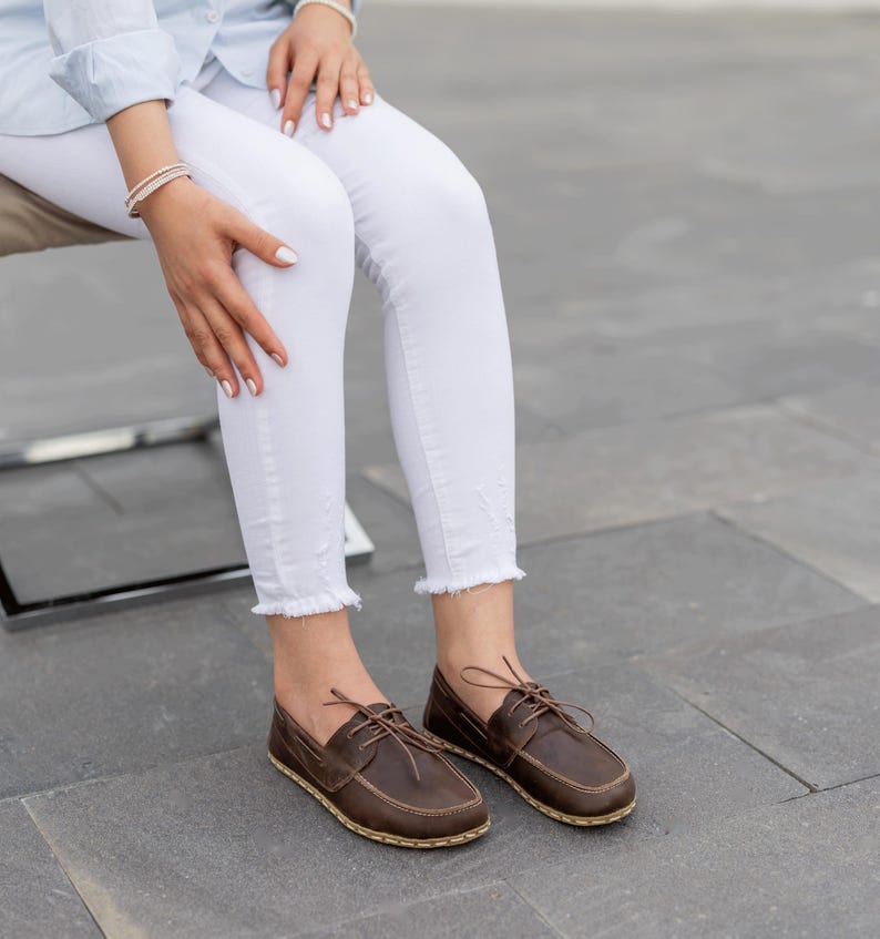 May include: Brown leather boat shoes with laces. The shoes have a stitched sole and are worn with white jeans. The shoes are a dark brown color.