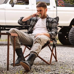 May include: A person seated in a folding chair, wearing a white t-shirt, gray pants, and black boots. They are drinking from a green cup. A black and white plaid shirt is draped over their shoulders. A white truck is in the background.