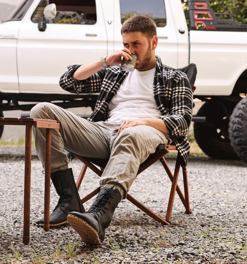 May include: A person seated in a folding chair, wearing a white t-shirt, gray pants, and black boots. They are drinking from a green cup. A black and white plaid shirt is draped over their shoulders. A white truck is in the background.